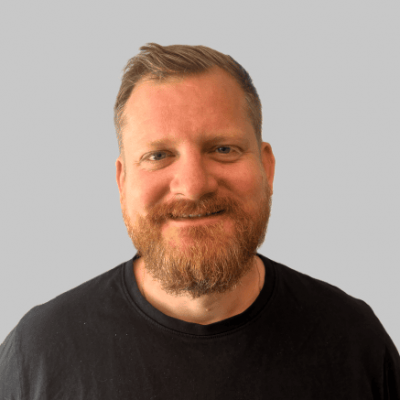 Portrett av en mann med kort, lyst hår og fullt skjegg, smilende, mot en nøytral grå bakgrunn, brukt i 'Om oss' siden for ElMeC.